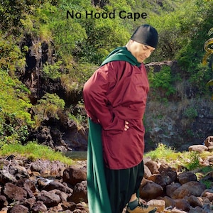 Może przedstawiać: Osoba w czerwono-zielonej pelerynie z kapturem i czarnym kapeluszem. Peleryna jest ściągnięta na głowę, a osoba patrzy w dół. Osoba stoi w skalistym terenie z zieloną roślinnością w tle. Tekst "No Hood Cape" jest napisany nad głową osoby.