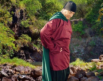 Peut inclure: Une personne portant une cape à capuche rouge et verte avec un chapeau noir. La cape est tirée sur la tête et la personne regarde vers le bas. La personne se tient dans une zone rocheuse avec une végétation verte en arrière-plan. Le texte "No Hood Cape" est écrit au-dessus de la tête de la personne.