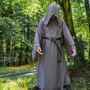 May include: A floor-length, solid grey hooded robe with long, wide sleeves. The garment is cinched at the waist with a black belt featuring a silver-toned metal ring buckle and a small black pouch attached to the side, creating a draped silhouette.