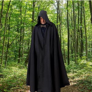 Puede incluir: Una capa negra con capucha de cuerpo entero que presenta una capucha estructurada y un cierre metálico decorativo en el cuello. La prenda es de color negro sólido, cae hasta el suelo y se lleva sobre una camisa oscura, creando un aspecto uniforme.