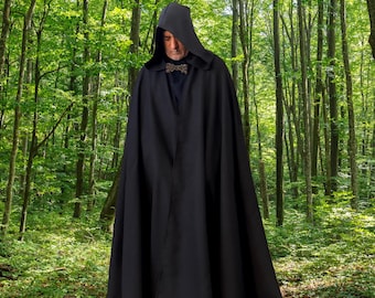 Peut inclure: Une cape à capuche noire pleine longueur dotée d'une capuche structurée et d'une attache métallique décorative au niveau du col. Le vêtement est entièrement noir, tombe jusqu'au sol et se porte sur une chemise sombre, créant un aspect uniforme.