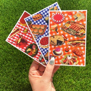 May include: Three colorful cards featuring picnic-themed illustrations. Each card showcases a different checkered pattern: red, white, and blue. The cards depict various picnic foods, including sandwiches, pies, and salads, creating a nostalgic, vintage aesthetic.