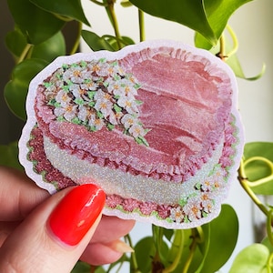 May include: A pink, glittery sticker in the shape of a cake, with white and pink floral accents. The sticker has a layered design with a ruffled edge and a sparkly, iridescent finish. The sticker is held by a hand with red nail polish.