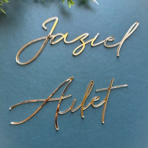 May include: Two gold-colored acrylic names, "Jaziel" and "Ailet", are displayed on a blue background. The names are written in a cursive script.