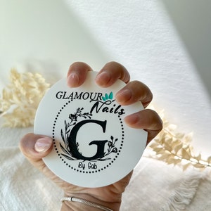 May include: A white, circular coaster featuring the text "GLAMOUR Nails by Gab" in black script, surrounded by a floral design. The coaster is held in a hand with manicured nails, and a silver bracelet is visible.