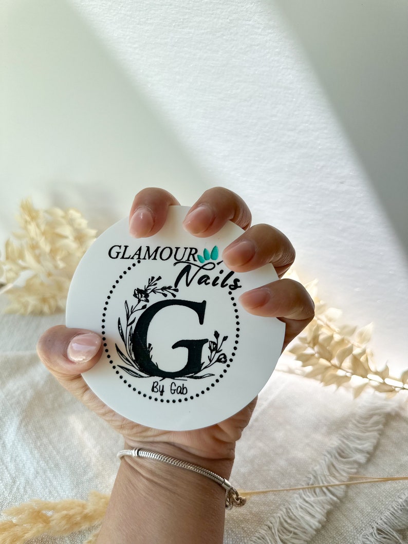 May include: A white, circular coaster featuring the text "GLAMOUR Nails by Gab" in black script, surrounded by a floral design. The coaster is held in a hand with manicured nails, and a silver bracelet is visible.
