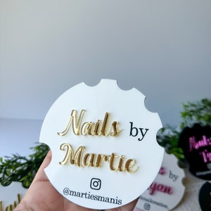 May include: A white, circular coaster with a scalloped edge. The coaster features gold lettering that reads "Nails by Martie" and an Instagram handle. Other coasters are visible in the background.