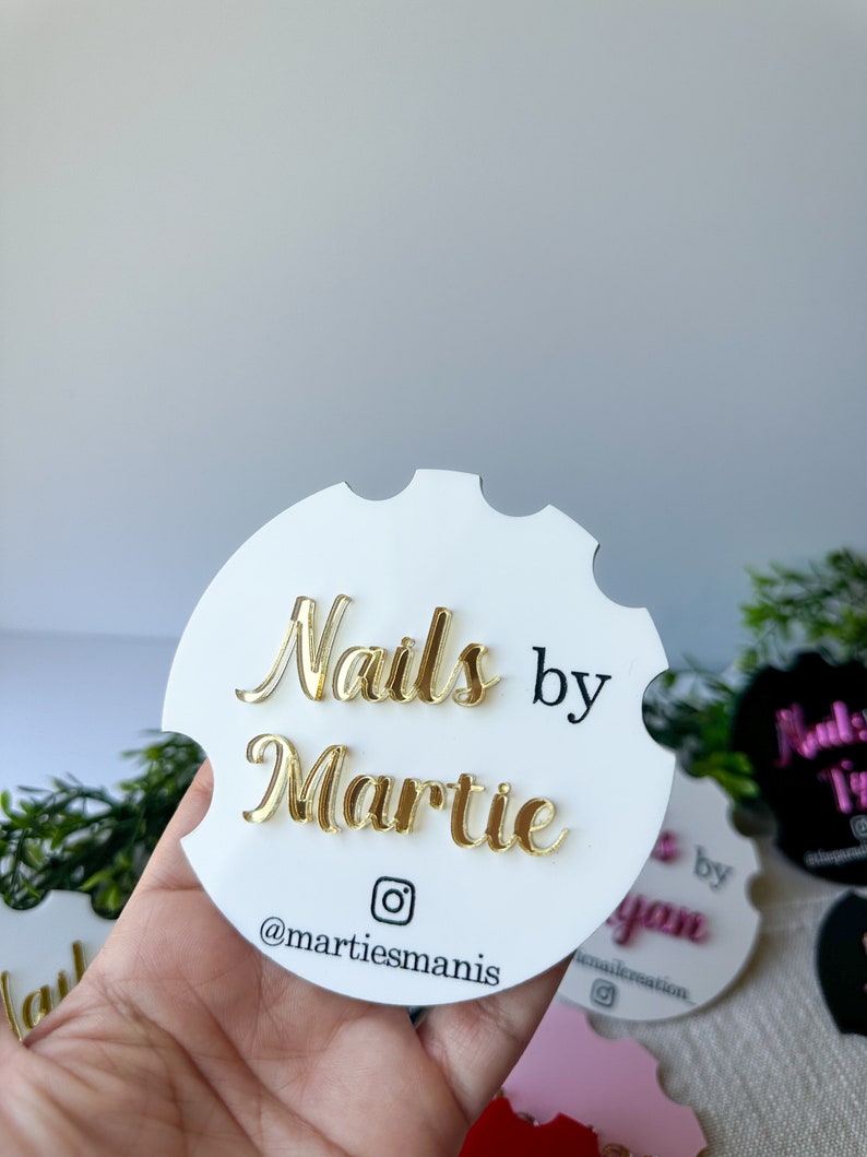May include: A white, circular coaster with a scalloped edge. The coaster features gold lettering that reads "Nails by Martie" and an Instagram handle. Other coasters are visible in the background.