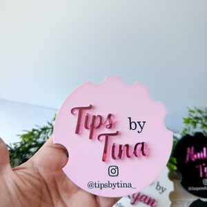 May include: A pink car coaster featuring the mirrored text "Tips by Tina" in a cursive font. The Instagram handle "@tipsbytina_" is displayed below. The coaster has a notched edge.