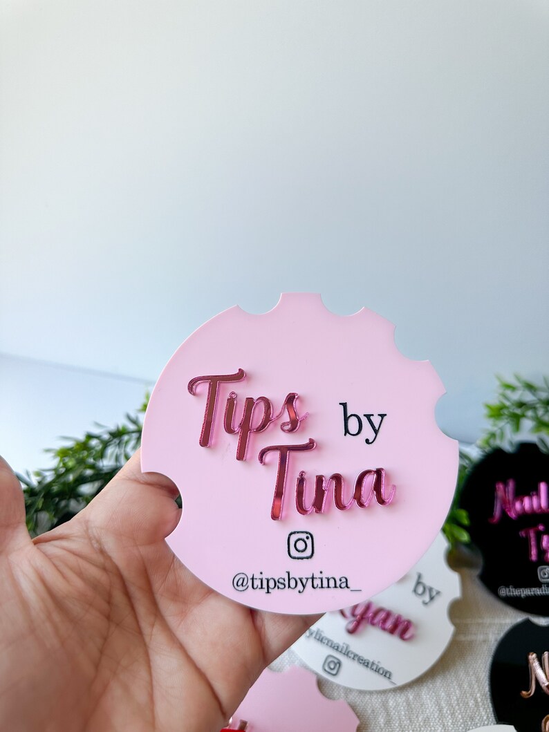 May include: A pink car coaster featuring the mirrored text "Tips by Tina" in a cursive font. The Instagram handle "@tipsbytina_" is displayed below. The coaster has a notched edge.