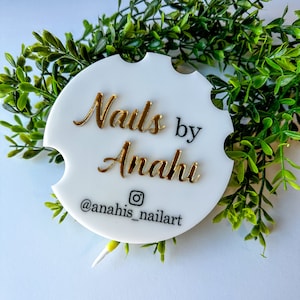 May include: White, circular car coaster with gold lettering reading "Nails by Anahi" and an Instagram handle. The coaster is surrounded by green foliage. A decorative item.