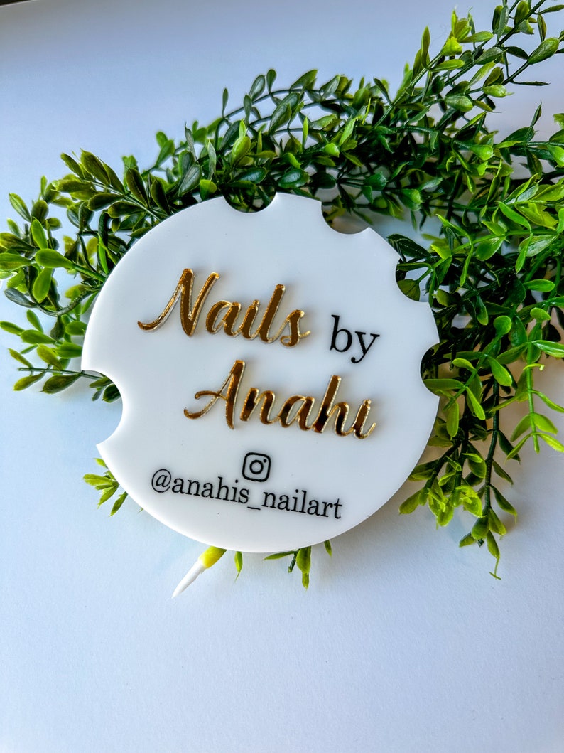 May include: White, circular car coaster with gold lettering reading "Nails by Anahi" and an Instagram handle. The coaster is surrounded by green foliage. A decorative item.