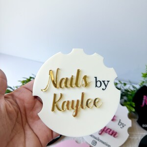 May include: A white car coaster with the text "Nails by Kaylee" in gold and black. The coaster has a scalloped edge and is held in a hand. Other coasters in pink, black, and red are visible.
