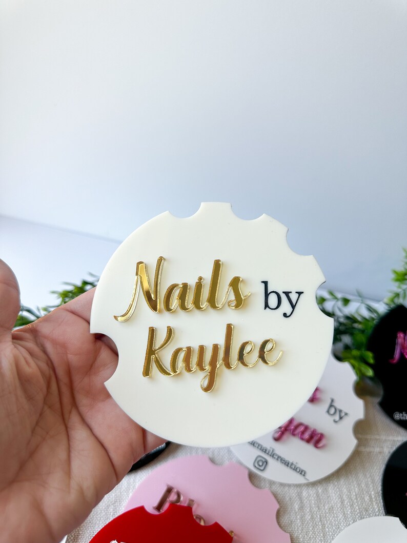 May include: A white car coaster with the text "Nails by Kaylee" in gold and black. The coaster has a scalloped edge and is held in a hand. Other coasters in pink, black, and red are visible.