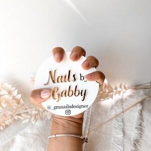 May include: A white circular sign with gold lettering that reads "Nails by Gabby" and the Instagram handle "@_gmnailsdesigner". The sign is held by a hand with manicured nails, against a soft, neutral background.