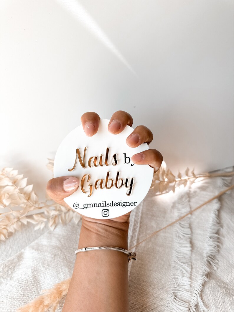 May include: A white circular sign with gold lettering that reads "Nails by Gabby" and the Instagram handle "@_gmnailsdesigner". The sign is held by a hand with manicured nails, against a soft, neutral background.