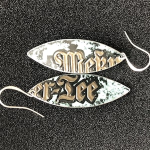 Może przedstawiać: Para srebrnych kolczyków o owalnym kształcie z efektem postarzenia. Każdy kolczyk zawiera napis "Rebel Tee" gotycką czcionką. Kolczyki mają srebrne haczyki.