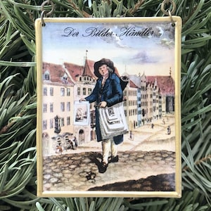 Puede incluir: Un adorno navideño de estilo vintage que presenta una pintura de un hombre vendiendo imágenes en una calle de la ciudad. El adorno es rectangular y tiene un marco dorado. El texto "Der Bilder-Handler" está escrito en la parte superior del adorno.