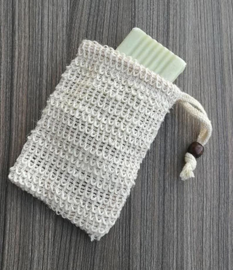 Puede incluir: Una bolsa de malla blanca con cierre de cord&oacute;n y una cuenta de madera. La bolsa est&aacute; llena de una barra de jab&oacute;n verde.