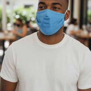 May include: A person wearing a light blue face mask and a white t-shirt. The mask covers the nose and mouth, secured by white elastic ear loops. The t-shirt is plain and has short sleeves.