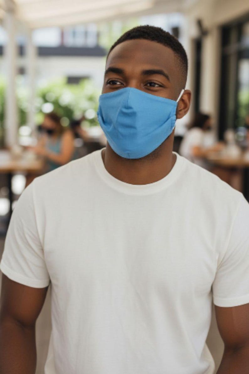 May include: A person wearing a light blue face mask and a white t-shirt. The mask covers the nose and mouth, secured by white elastic ear loops. The t-shirt is plain and has short sleeves.