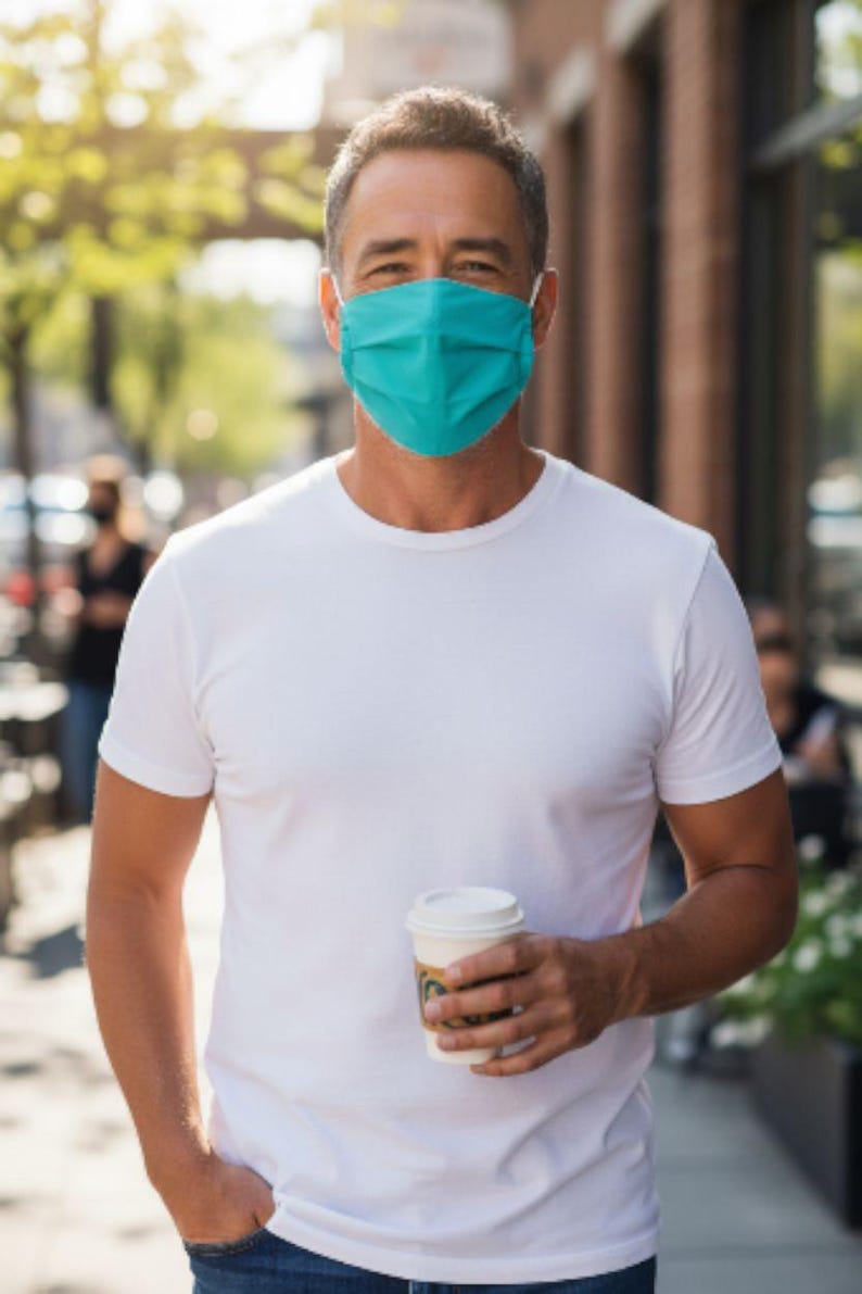 May include: A person wearing a teal face mask and a white t-shirt, holding a takeaway coffee cup. The person is outdoors on a sunny day. The background includes a brick building and foliage.