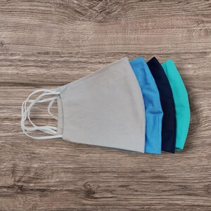 May include: Five fabric face masks in shades of blue and grey, arranged on a wooden surface. The masks have white elastic ear loops. The colours include light grey, light blue, medium blue, navy, and teal. The masks are made of fabric.