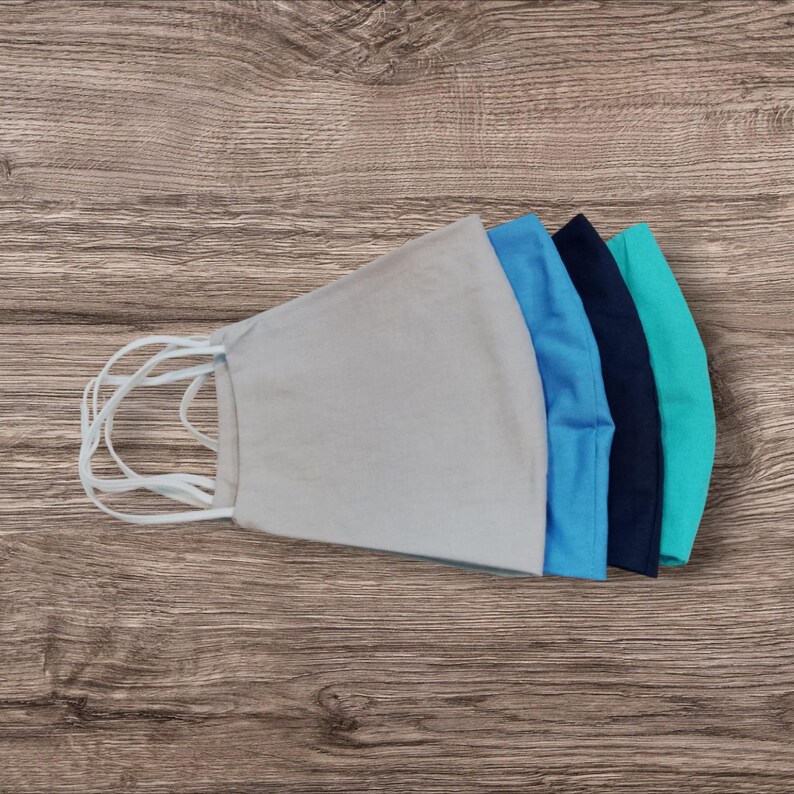 May include: Five fabric face masks in shades of blue and grey, arranged on a wooden surface. The masks have white elastic ear loops. The colours include light grey, light blue, medium blue, navy, and teal. The masks are made of fabric.