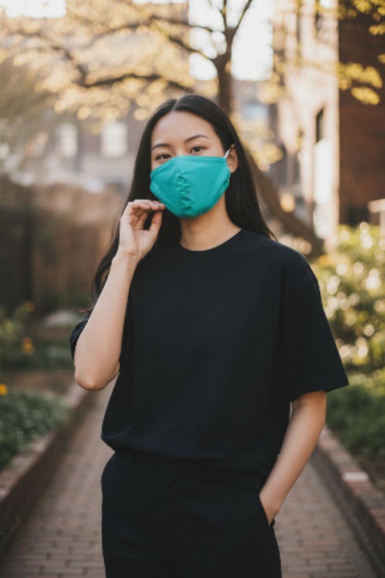 May include: A teal face mask with a stitched design is worn by a person wearing a black t-shirt and black trousers. The background shows a brick pathway and trees.