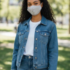 May include: A person wearing a light grey face mask, a denim jacket, a white t-shirt, and blue jeans. The jacket has button closures and pockets. The background is a blurred outdoor scene with trees.