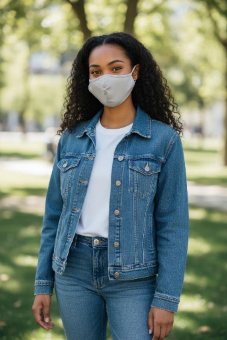 May include: A person wearing a light grey face mask, a denim jacket, a white t-shirt, and blue jeans. The jacket has button closures and pockets. The background is a blurred outdoor scene with trees.