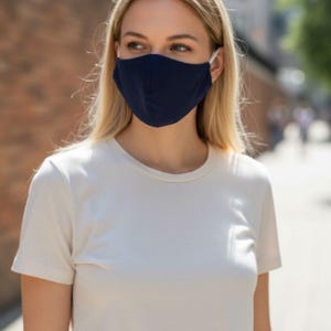 May include: A person wearing a navy blue face mask with white ear loops and a white t-shirt. The background features a brick wall and a blurred street scene.