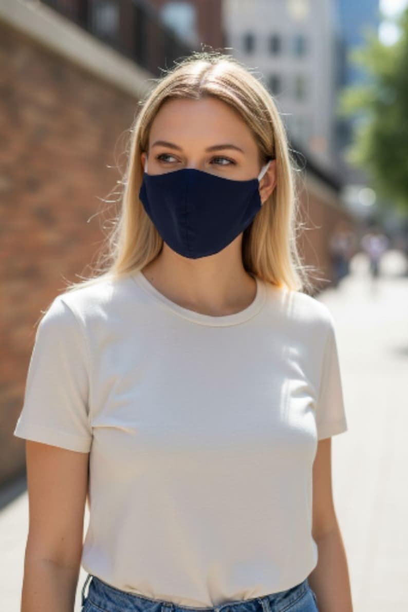 May include: A person wearing a navy blue face mask with white ear loops and a white t-shirt. The background features a brick wall and a blurred street scene.
