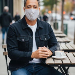 May include: A man wearing a light grey face mask, white t-shirt, dark blue denim jacket, and blue jeans sits at an outdoor cafe table. A white coffee cup and saucer are on the table. The background shows a city street.
