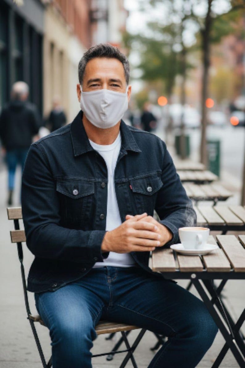 May include: A man wearing a light grey face mask, white t-shirt, dark blue denim jacket, and blue jeans sits at an outdoor cafe table. A white coffee cup and saucer are on the table. The background shows a city street.