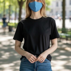 May include: A person wearing a blue face mask, black t-shirt, and blue jeans. The person is standing outdoors in front of a blurred background of trees and a walkway.