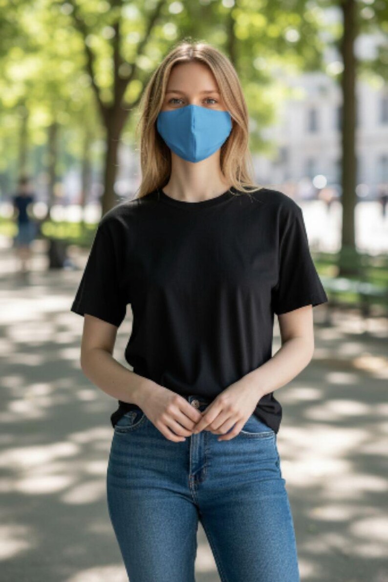 May include: A person wearing a blue face mask, black t-shirt, and blue jeans. The person is standing outdoors in front of a blurred background of trees and a walkway.