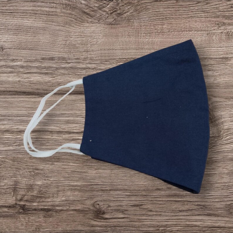 May include: A navy blue face covering with white elastic ear loops. The mask is made of a solid, dark blue fabric and is designed to cover the nose and mouth. The mask is laying on a wooden surface.