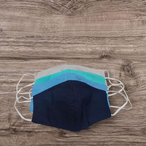 May include: Five face masks in a stack, featuring a colour palette of blues and grey, with white elastic ear loops. The masks are arranged on a wooden surface, with the colours ranging from navy to light blue and grey.