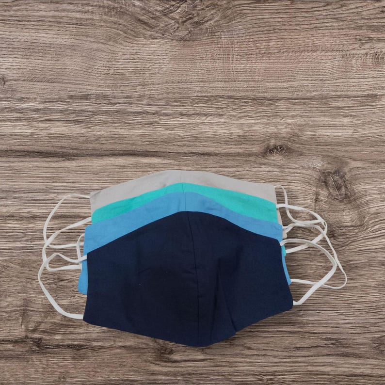 May include: Five face masks in a stack, featuring a colour palette of blues and grey, with white elastic ear loops. The masks are arranged on a wooden surface, with the colours ranging from navy to light blue and grey.