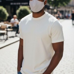 May include: A person wearing a white t-shirt, blue jeans, and a light grey face mask. The person is standing outdoors on a sunny day with their hands in their pockets.