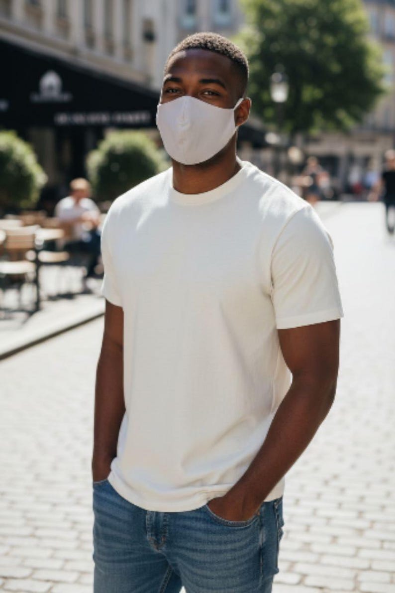 May include: A person wearing a white t-shirt, blue jeans, and a light grey face mask. The person is standing outdoors on a sunny day with their hands in their pockets.