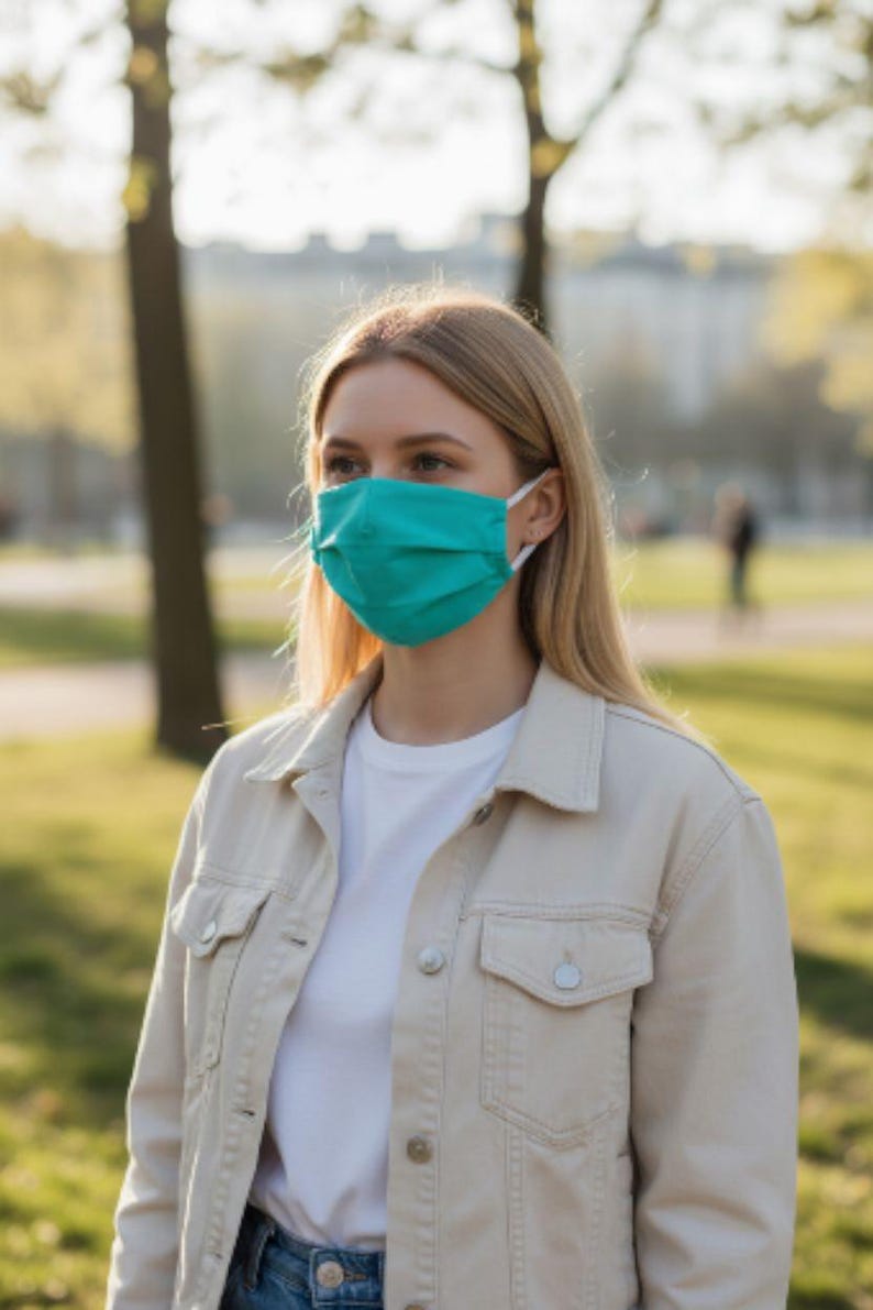 May include: A teal face mask with white ear loops. The mask is worn by a person wearing a white t-shirt and a light beige denim jacket. The background is a park with trees and sunlight.