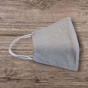 May include: A pale grey face mask with white elastic ear loops. The mask is made of a soft fabric and is designed to cover the nose and mouth. The mask is shown on a wooden surface.
