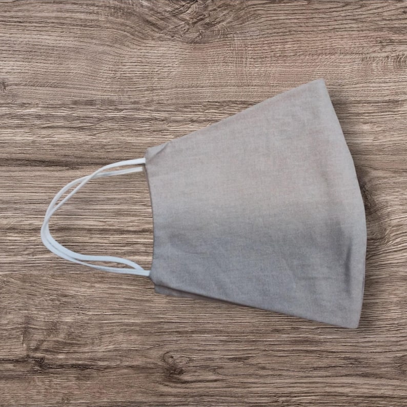 May include: A pale grey face mask with white elastic ear loops. The mask is made of a soft fabric and is designed to cover the nose and mouth. The mask is shown on a wooden surface.