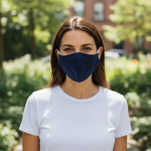 May include: A navy blue face mask with white ear loops. The mask is worn by a person wearing a white t-shirt. The background features a blurred outdoor scene with trees and a building.