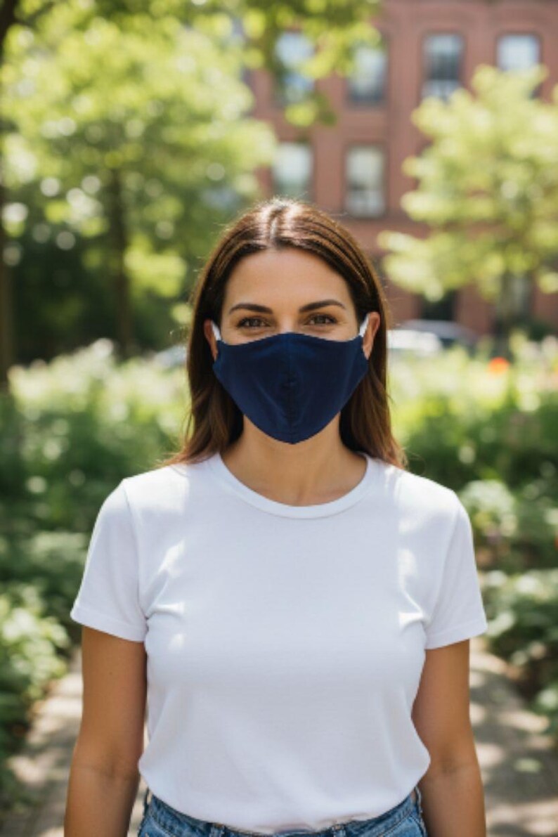 May include: A navy blue face mask with white ear loops. The mask is worn by a person wearing a white t-shirt. The background features a blurred outdoor scene with trees and a building.