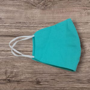 May include: A teal face mask with white elastic ear loops. The mask is made of a soft fabric and is designed to cover the nose and mouth. The mask is shown on a wooden surface.