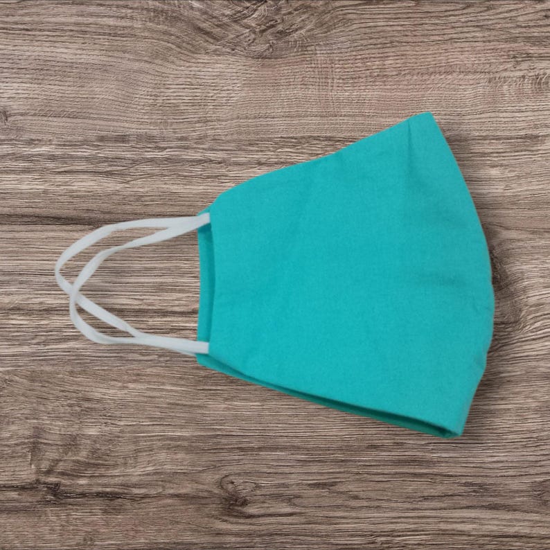 May include: A teal face mask with white elastic ear loops. The mask is made of a soft fabric and is designed to cover the nose and mouth. The mask is shown on a wooden surface.