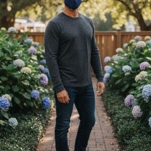 May include: A person wearing a navy blue face mask, dark grey long-sleeved top, and dark blue jeans. They are standing on a brick path lined with hydrangeas. Brown leather shoes complete the outfit.